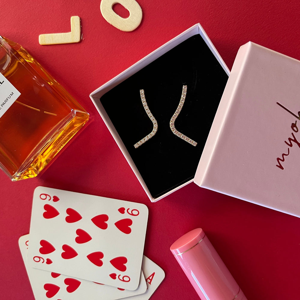 Packaging rosa quarso di MyOh su un tavolo di gioco rosso, un profumo chanel e orecchini zirconi della collezione di gioielli Myoh. Idee regalo per lei