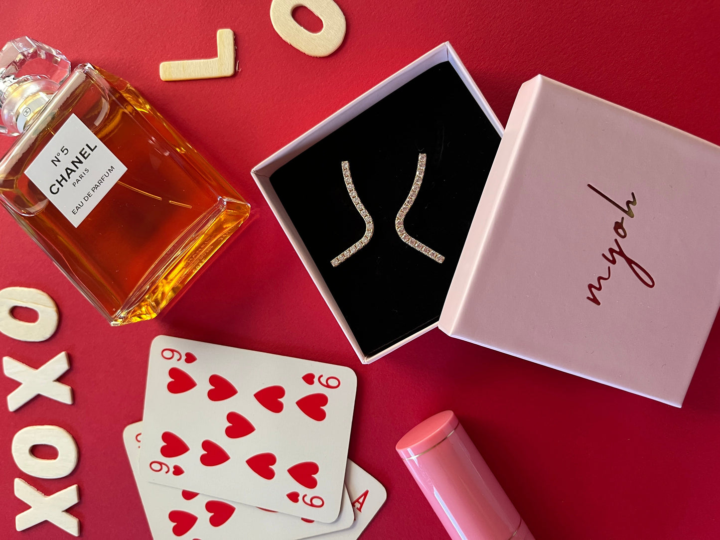 Packaging rosa quarso di MyOh su un tavolo di gioco rosso, un profumo chanel e orecchini zirconi della collezione di gioielli Myoh. Idee regalo per lei